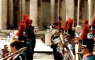 Colour photograph showing the brass section of a parade band marching past the Ploegsteert Memorial to the Missing at the Royal Berkshire Cemetery Extension.