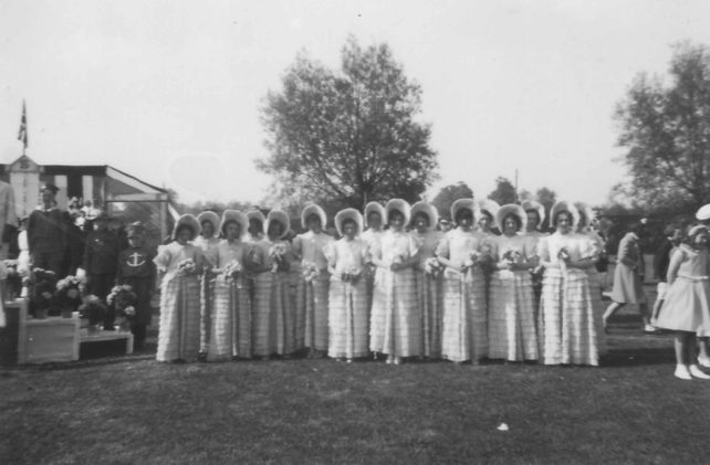 A group of 16 women dressed in identical bonnets and dresses with frilly skirts and bouquets
