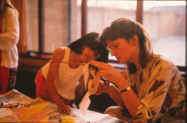 A girl watching a woman cutting out a mask