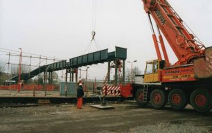 Crane holding a station footbridge span as fitters bolt it to uprights