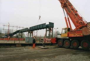 Crane holding a station footbridge span as fitters bolt it to uprights